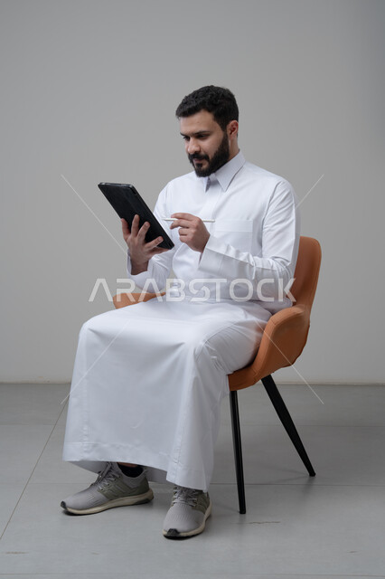 Browsing social media, using technology and modern technology, portrait of a Saudi Arabian Gulf man wearing traditional thobe sitting on a comfortable chair working on a tablet, chatting with family and friends via tablet, gray background