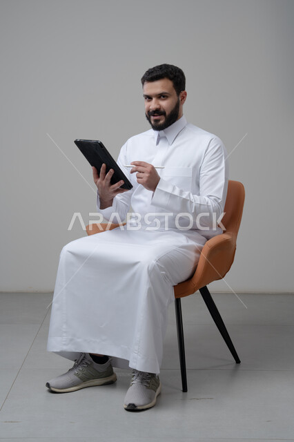 Browsing social media, using technology and modern technology, portrait of a Saudi Arabian Gulf man wearing traditional thobe sitting on a comfortable chair working on a tablet, chatting with family and friends via tablet, gray background