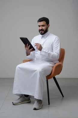 Browsing social media, using technology and modern technology, portrait of a Saudi Arabian Gulf man wearing traditional thobe sitting on a comfortable chair working on a tablet, chatting with family and friends via tablet, gray background
