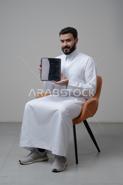 عرض شاشة سوداء فارغة عبر التابلت، تصفح مواقع التواصل الاجتماعي، استخدام التكنولوجيا والتقنية الحديثة, بورتريه لرجل عربي خليجي سعودي يرتدي الثوب التقليدي يجلس على كرسي مريح يعمل على الجهاز اللوحي، خلفية رمادية