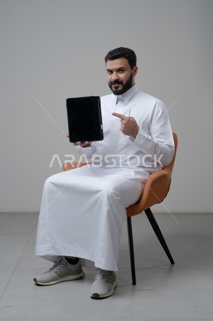 عرض شاشة سوداء فارغة عبر التابلت، تصفح مواقع التواصل الاجتماعي، استخدام التكنولوجيا والتقنية الحديثة, بورتريه لرجل عربي خليجي سعودي يرتدي الثوب التقليدي يجلس على كرسي مريح يمسك بيده الجهاز اللوحي يشير اليه باصبع السبابة، خلفية رمادية