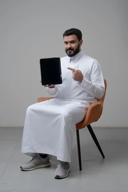 عرض شاشة سوداء فارغة عبر التابلت، تصفح مواقع التواصل الاجتماعي، استخدام التكنولوجيا والتقنية الحديثة, بورتريه لرجل عربي خليجي سعودي يرتدي الثوب التقليدي يجلس على كرسي مريح يمسك بيده الجهاز اللوحي يشير اليه باصبع السبابة، خلفية رمادية