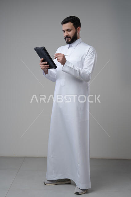 Using modern technology and techniques, browsing social media, portrait of a Saudi Arabian Gulf man wearing traditional thobe working on a tablet, chatting with family and friends via tablet, full-length body image, gray background