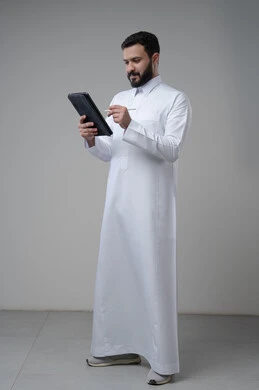 Using modern technology and techniques, browsing social media, portrait of a Saudi Arabian Gulf man wearing traditional thobe working on a tablet, chatting with family and friends via tablet, full-length body image, gray background