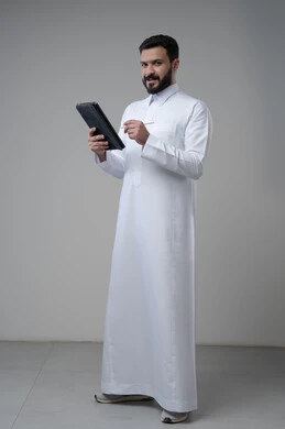 Using modern technology and techniques, browsing social media, portrait of a Saudi Arabian Gulf man wearing traditional thobe working on a tablet, chatting with family and friends via tablet, full-length body image, gray background