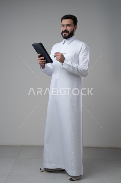 Using modern technology and techniques, browsing social media, portrait of a Saudi Arabian Gulf man wearing traditional thobe working on a tablet, chatting with family and friends via tablet, full-length body image, gray background