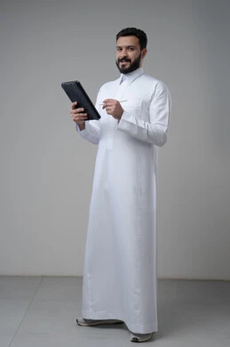 Using modern technology and techniques, browsing social media, portrait of a Saudi Arabian Gulf man wearing traditional thobe working on a tablet, chatting with family and friends via tablet, full-length body image, gray background