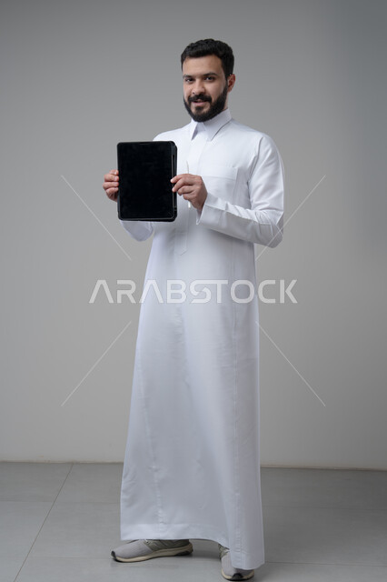 Blank black screen on tablet, browsing social media, using technology and modern technology, portrait of a Saudi Arabian Gulf man wearing traditional thobe holding a tablet in his hand looking at the camera with an expression of pleasure, full body image, gray background