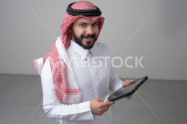 Using modern technology and techniques, browsing social media, close-up portrait of a Saudi Arabian Gulf man wearing traditional thobe and shemagh working on a tablet, chatting with family and friends via tablet, looking at the camera with a happy expression, gray background