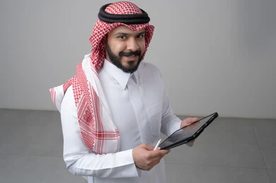 Using modern technology and techniques, browsing social media, close-up portrait of a Saudi Arabian Gulf man wearing traditional thobe and shemagh working on a tablet, chatting with family and friends via tablet, looking at the camera with a happy expression, gray background