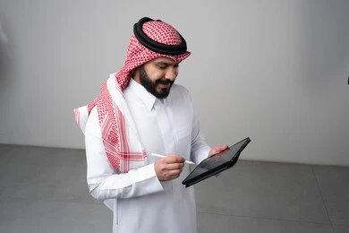 استخدام التكنولوجيا والتقنية الحديثة, تصفح مواقع التواصل الاجتماعي، بورتريه مقرب لرجل عربي خليجي سعودي يرتدي الثوب التقليدي والشماغ يعمل على التابليت، الدردشة مع الأهل والأصدقاء عبر الجهاز اللوحي بتعابير الاندماج, خلفية رمادية