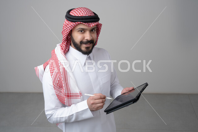 استخدام التكنولوجيا والتقنية الحديثة, تصفح مواقع التواصل الاجتماعي، بورتريه مقرب لرجل عربي خليجي سعودي يرتدي الثوب التقليدي والشماغ يعمل على التابليت، الدردشة مع الأهل والأصدقاء عبر الجهاز اللوحي، النظر الى الكاميرا بتعابير السرور, خلفية رمادية