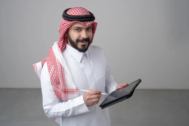 Using modern technology and techniques, browsing social media, close-up portrait of a Saudi Arabian Gulf man wearing traditional thobe and shemagh working on a tablet, chatting with family and friends via tablet, looking at the camera with a happy expression, gray background
