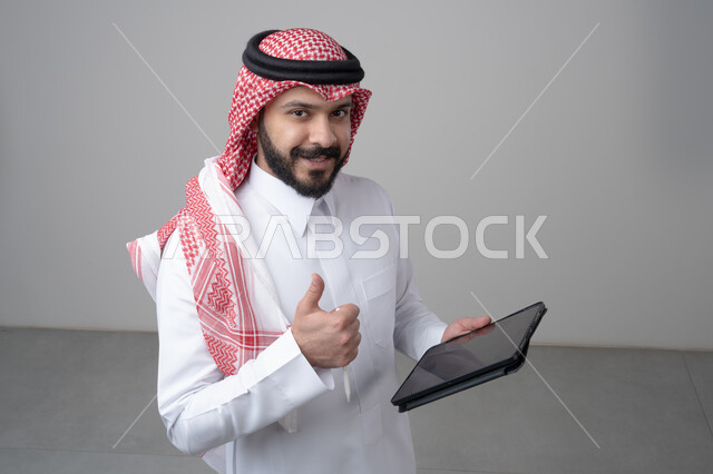 Using modern technology and techniques, browsing social media, close-up portrait of a Saudi Arabian Gulf man wearing traditional thobe and shemagh working on a tablet, chatting with family and friends via tablet, looking at the camera with a happy expression, gray background