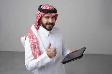Using modern technology and techniques, browsing social media, close-up portrait of a Saudi Arabian Gulf man wearing traditional thobe and shemagh working on a tablet, chatting with family and friends via tablet, looking at the camera with a happy expression, gray background