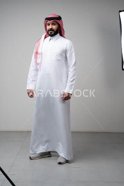  الاعتناء والاهتمام بالمظهر الخارجي، الوقوف بجدية وحزم، مفهوم الاناقة والرجولة، بورتريه لشاب عربي خليجي سعودي يرتدي الشماغ والثوب التقليدي يقف باستقامة ينظر الى الكاميرا بتعابير الثقة بالنفس، الجسم بالطول الكامل، خلفية رمادية