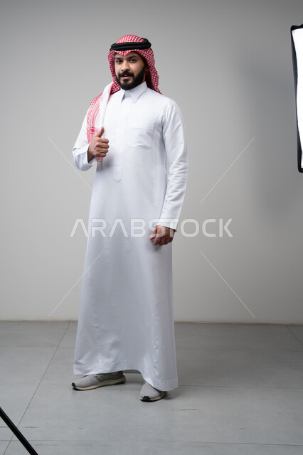 Using modern technology and techniques, browsing social media. Close-up portrait of a Saudi Arabian Gulf man wearing a traditional thobe and keffiyeh working on a tablet. Chatting with family and friends on the tablet with an expression of engagement. Gray background.  Thumbs up, looking at the camera with expressions of admiration and satisfaction. Portrait of a Saudi Arabian Gulf man wearing a keffiyeh and traditional thobe looking at the camera with gestures of acceptance and approval. Full-l