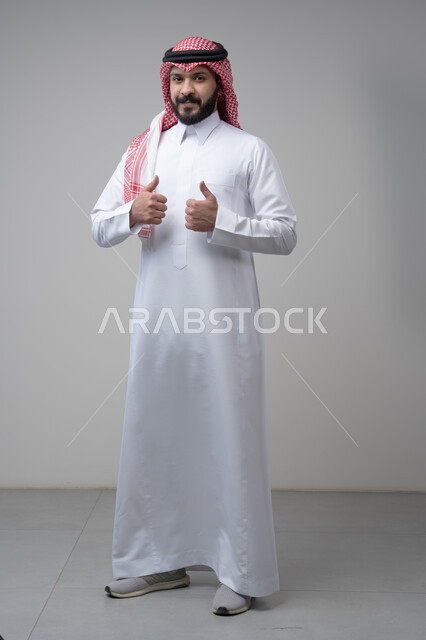 Using modern technology and techniques, browsing social media. Close-up portrait of a Saudi Arabian Gulf man wearing a traditional thobe and keffiyeh working on a tablet. Chatting with family and friends on the tablet with an expression of engagement. Gray background.  Thumbs up, looking at the camera with expressions of admiration and satisfaction. Portrait of a Saudi Arabian Gulf man wearing a keffiyeh and traditional thobe looking at the camera with gestures of acceptance and approval. Full-l