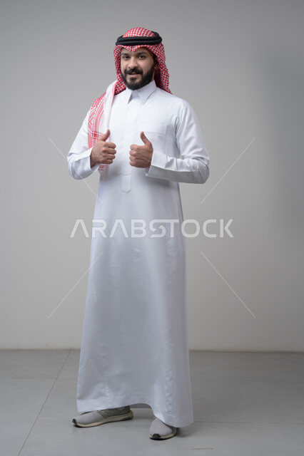 Using modern technology and techniques, browsing social media. Close-up portrait of a Saudi Arabian Gulf man wearing a traditional thobe and keffiyeh working on a tablet. Chatting with family and friends on the tablet with an expression of engagement. Gray background.  Thumbs up, looking at the camera with expressions of admiration and satisfaction. Portrait of a Saudi Arabian Gulf man wearing a keffiyeh and traditional thobe looking at the camera with gestures of acceptance and approval. Full-l