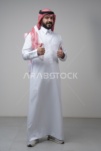 رفع اصابع الابهام الى الاعلى، النظر الى الكاميرا  بتعابير الاعجاب والرضا، بورتريه لرجل عربي خليجي سعودي يرتدي الشماغ والثوب التقليدي ينظر الى الكاميرا بايماءات القبول والموافقة، صورة الجسم بالطول الكامل، خلفية رمادية