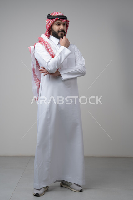 Looking forward and planning for the future, solving problems and making decisions, portrait of a Saudi Arabian Gulf man wearing a shemagh and a Saudi thobe, putting his hand on his chin with gestures of thinking and absent-mindedness, the concept of scrutiny and concentration, full-length body, gray background