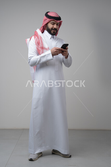 Gestures and expressions of integration and concentration while working on the mobile phone, use of technology and technology in daily life, portrait of a Saudi Arabian Gulf man wearing a traditional shemagh and thobe holding a mobile phone in his hand, full body image, gray background