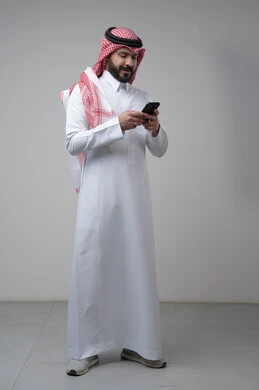 Gestures and expressions of integration and concentration while working on the mobile phone, use of technology and technology in daily life, portrait of a Saudi Arabian Gulf man wearing a traditional shemagh and thobe holding a mobile phone in his hand, full body image, gray background