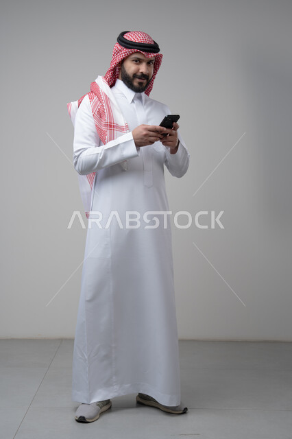ايماءات وتعابير الاندماج والتركيز بالعمل على الجوال، استخدام التقنية والتكنولوجيا في الحياة اليومية، بورتريه لرجل عربي خليجي سعودي يرتدي الشماغ والثوب التقليدي يمسك بيده الهاتف المحمول، صورة الجسم بالطول الكامل، خلفية رمادية