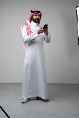 Gestures and expressions of integration and concentration while working on the mobile phone, use of technology and technology in daily life, portrait of a Saudi Arabian Gulf man wearing a traditional shemagh and thobe holding a mobile phone in his hand, full body image, gray background