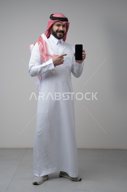 Blank black screen view on mobile, use of technology in daily life, portrait of a Saudi Arabian Gulf man wearing a traditional shemagh and thobe pointing at a mobile phone, full body image, gray background