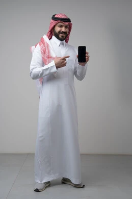 عرض شاشة سوداء فارغة عبر الجوال، استخدام التقنية والتكنولوجيا في الحياة اليومية، بورتريه لرجل عربي خليجي سعودي يرتدي الشماغ والثوب التقليدي يقوم بالاشارة الى الهاتف المحمول، صورة الجسم بالطول الكامل، خلفية رمادية