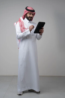 استخدام التكنولوجيا والتقنية الحديثة, تصفح مواقع التواصل الاجتماعي، بورتريه لرجل عربي خليجي سعودي يرتدي الشماغ والثوب التقليدي يعمل على التابليت، الدردشة مع الأهل والأصدقاء عبر الجهاز اللوحي، صورة الجسم بالطول الكامل, خلفية رمادية