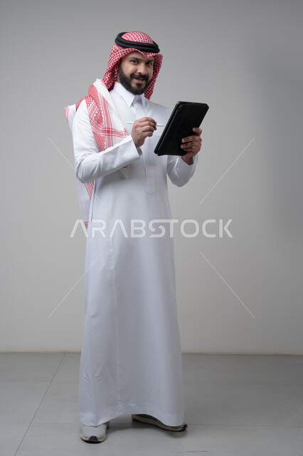 استخدام التكنولوجيا والتقنية الحديثة, تصفح مواقع التواصل الاجتماعي، بورتريه لرجل عربي خليجي سعودي يرتدي الشماغ والثوب التقليدي يعمل على التابليت، الدردشة مع الأهل والأصدقاء عبر الجهاز اللوحي، صورة الجسم بالطول الكامل, خلفية رمادية