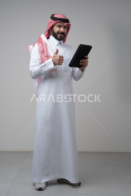 رفع اصبع الابهام لاعلى بتعابير القبول والاعجاب، استخدام التكنولوجيا والتقنية الحديثة، بورتريه لرجل عربي خليجي سعودي يرتدي الشماغ والثوب التقليدي يعمل على التابليت، الدردشة مع الأهل والأصدقاء عبر الجهاز اللوحي، صورة الجسم بالطول الكامل, خلفية رمادية