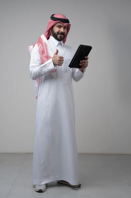 رفع اصبع الابهام لاعلى بتعابير القبول والاعجاب، استخدام التكنولوجيا والتقنية الحديثة، بورتريه لرجل عربي خليجي سعودي يرتدي الشماغ والثوب التقليدي يعمل على التابليت، الدردشة مع الأهل والأصدقاء عبر الجهاز اللوحي، صورة الجسم بالطول الكامل, خلفية رمادية