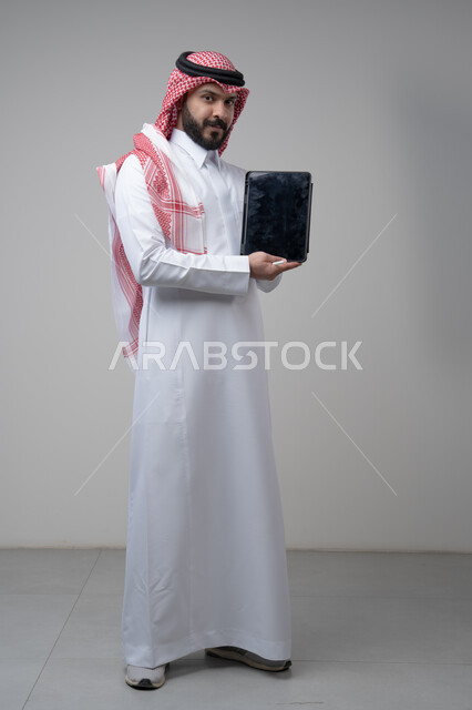 عرض شاشة سوداء فارغة عبر التابلت، تصفح مواقع التواصل الاجتماعي، استخدام التكنولوجيا والتقنية الحديثة, بورتريه لرجل عربي خليجي سعودي يرتدي الشماغ والثوب التقليدي يمسك بيده الجهاز اللوحي ينظر الى الكاميرا بتعابير السرور، صورة الجسم بالطول الكامل، خلفية رمادية