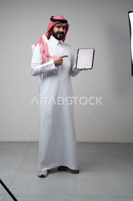 Using modern technology and techniques, browsing social media, close-up portrait of a Saudi Arabian Gulf man wearing a traditional keffiyeh and thobe, working on a tablet, chatting with family and friends via the tablet with expressions of integration, gray background