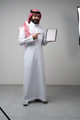 Using modern technology and techniques, browsing social media, close-up portrait of a Saudi Arabian Gulf man wearing a traditional keffiyeh and thobe, working on a tablet, chatting with family and friends via the tablet with expressions of integration, gray background