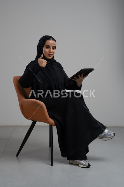 رفع اصبع الابهام لاعلى بتعابير الاعجاب والقبول، انجاز وادارة المهام باستخدام جهاز تقني حديث ومتطور, العمل عن بعد، بورتريه لإمرأة عربية خليجية سعودية مبتسمة ترتدي الحجاب والعباءة السوداء تجلس على الكرسي و تعمل على جهاز التابلت, خلفية رمادية
