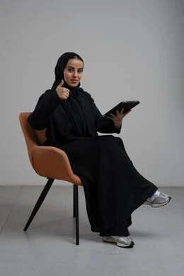 رفع اصبع الابهام لاعلى بتعابير الاعجاب والقبول، انجاز وادارة المهام باستخدام جهاز تقني حديث ومتطور, العمل عن بعد، بورتريه لإمرأة عربية خليجية سعودية مبتسمة ترتدي الحجاب والعباءة السوداء تجلس على الكرسي و تعمل على جهاز التابلت, خلفية رمادية