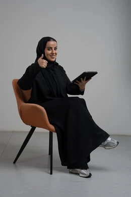 رفع اصبع الابهام لاعلى بتعابير الاعجاب والقبول، انجاز وادارة المهام باستخدام جهاز تقني حديث ومتطور, العمل عن بعد، بورتريه لإمرأة عربية خليجية سعودية مبتسمة ترتدي الحجاب والعباءة السوداء تجلس على الكرسي و تعمل على جهاز التابلت, خلفية رمادية