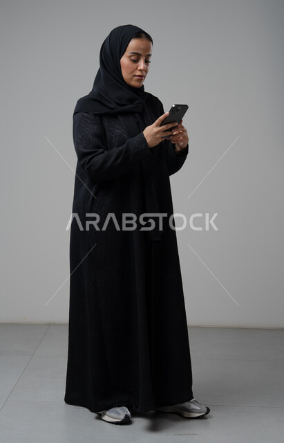استخدام جهاز تقني حديث، بورتريه لإمرأة عربية خليجية سعودية ترتدي الحجاب والعباءة السوداء تقوم بالعمل على الهاتف المحمول بتعابير السرور والاندماج، صورة الجسم بالطول الكامل، خلفية رمادية