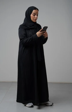 استخدام جهاز تقني حديث، بورتريه لإمرأة عربية خليجية سعودية ترتدي الحجاب والعباءة السوداء تقوم بالعمل على الهاتف المحمول بتعابير السرور والاندماج، صورة الجسم بالطول الكامل، خلفية رمادية