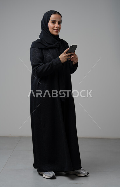 استخدام جهاز تقني حديث، بورتريه لإمرأة عربية خليجية سعودية ترتدي الحجاب والعباءة السوداء تقوم بالعمل على الهاتف المحمول بتعابير السرور والاندماج، صورة الجسم بالطول الكامل، خلفية رمادية