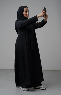 Documenting beautiful moments, taking selfies using a mobile phone, portrait of a Saudi Arabian Gulf woman wearing a hijab and a black abaya, holding a mobile phone with an expression of pleasure and happiness, using a modern technical device, full body, gray background