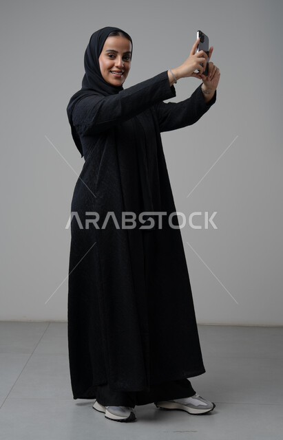 توثيق اللحظات الجميلة، التقاط صور سيلفي باستخدام الهاتف المحمول، بورتريه لإمرأة عربية خليجية سعودية ترتدي الحجاب والعباءة السوداء تحمل بيدها الجوال بتعابير السرور والسعادة، استخدام جهاز تقني حديث، الجسم بالطول الكامل، خلفية رمادية