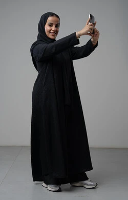 Documenting beautiful moments, taking selfies using a mobile phone, portrait of a Saudi Arabian Gulf woman wearing a hijab and a black abaya, holding a mobile phone with an expression of pleasure and happiness, using a modern technical device, full body, gray background