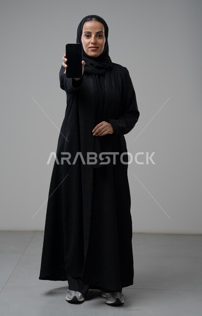 عرض شاشة سوداء فارغة عبر الهاتف المحمول، بورتريه لإمرأة عربية خليجية سعودية ترتدي الحجاب والعباءة السوداء تحمل بيدها الجوال بتعابير السرور والسعادة، استخدام جهاز تقني حديث، الجسم بالطول الكامل، خلفية رمادية