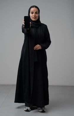 عرض شاشة سوداء فارغة عبر الهاتف المحمول، بورتريه لإمرأة عربية خليجية سعودية ترتدي الحجاب والعباءة السوداء تحمل بيدها الجوال بتعابير السرور والسعادة، استخدام جهاز تقني حديث، الجسم بالطول الكامل، خلفية رمادية