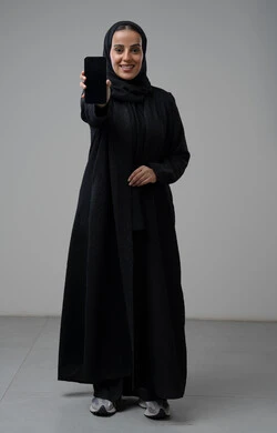 عرض شاشة سوداء فارغة عبر الهاتف المحمول، بورتريه لإمرأة عربية خليجية سعودية ترتدي الحجاب والعباءة السوداء تحمل بيدها الجوال بتعابير السرور والسعادة، استخدام جهاز تقني حديث، الجسم بالطول الكامل، خلفية رمادية